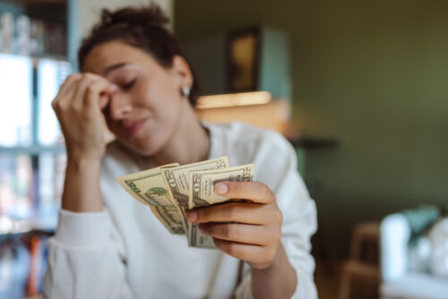 A worried young woman holding money while sitting at the table in the living room