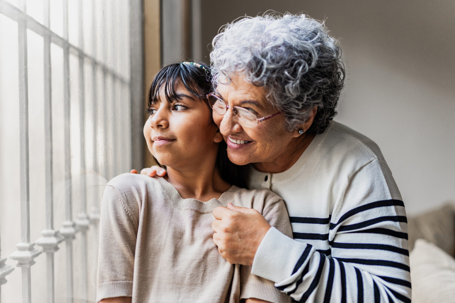 Grandmother and granddaughter looking through the window and contemplating at home